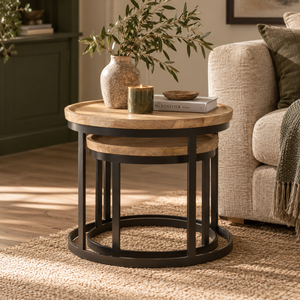 A round nesting side table with a light oak wood top and black metal frame, styled in a cosy living room next to a beige sofa with a vase and candle on top.