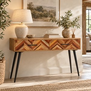 A mid-century modern wooden console table with chevron-patterned wood drawer fronts and black tapered legs, styled in a warm living room setting with a lamp, books, and plants.