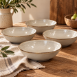 The Gable Collection Set of Four Stoneware Cereal Bowls, four soft beige bowls with brown rims arranged on a rustic wooden table with a linen cloth.