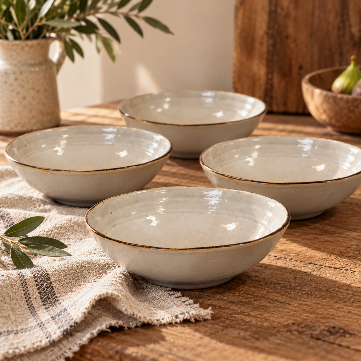 The Gable Collection Set of Four Stoneware Cereal Bowls, four soft beige bowls with brown rims arranged on a rustic wooden table with a linen cloth.