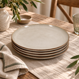 The Gable Collection Set of Four Stoneware Dinner Plates, stacked on a linen runner on a rustic wooden table with olive branches in a speckled vase.