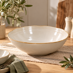 The Gable Collection Stoneware Salad Bowl, an empty soft beige serving bowl with a gently wavy brown rim, on a wooden dining table.