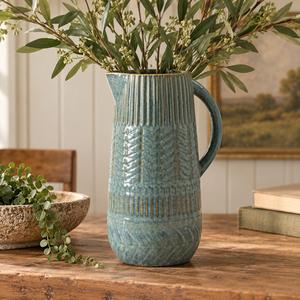 Decorative blue ceramic jug with textured detailing, styled with olive branches on a rustic wooden table in a warm country-style interior.