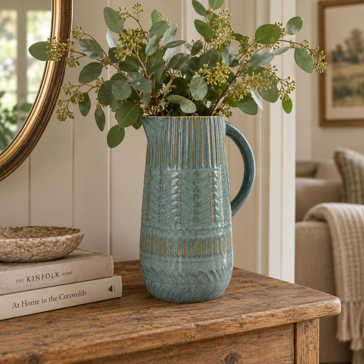 A tall blue-green decorative ceramic jug with textured detailing, styled with eucalyptus branches on a rustic wooden console in a warm country-style interior.