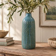 Tall blue ceramic vase with raised vertical detailing, styled with leafy branches on a rustic wooden table in a warm, neutral interior.
