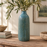 Tall blue ceramic vase with raised vertical detailing, styled with leafy branches on a rustic wooden table in a warm, neutral interior.