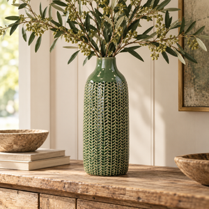 Tall deep green textured ceramic vase with olive branches displayed on a rustic wooden console in a warm, neutral country-style interior.
