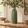 Tall deep green textured ceramic vase with olive branches displayed on a rustic wooden console in a warm, neutral country-style interior.