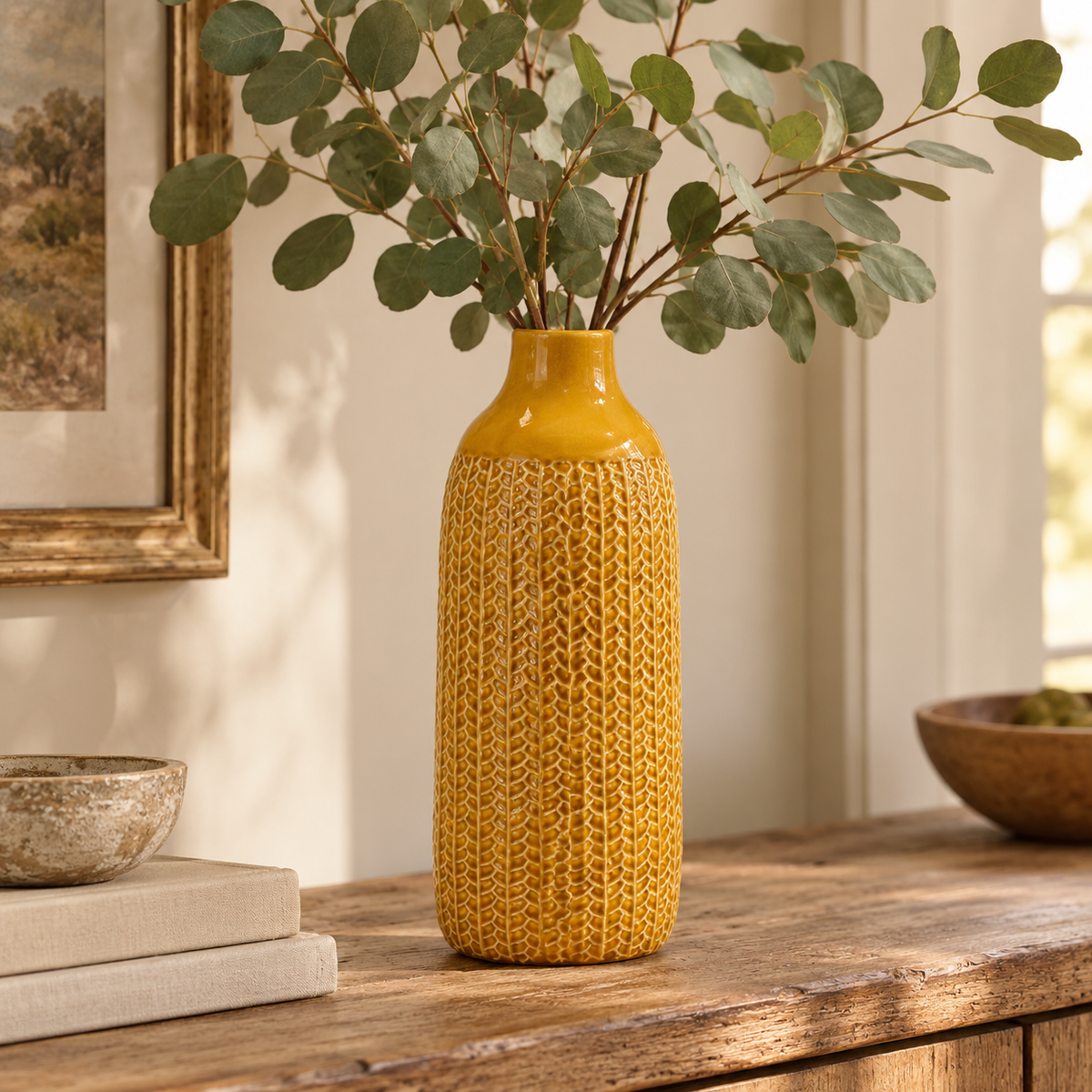 Mustard yellow ceramic vase with an intricate raised leaf-like pattern, styled with eucalyptus stems on a rustic wooden console table in a warm, neutral country-style interior.