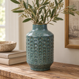 Textured teal ceramic vase with an embossed arch pattern, displayed on a rustic wooden table with olive branches, neutral books and a stone bowl in a warm, natural interior