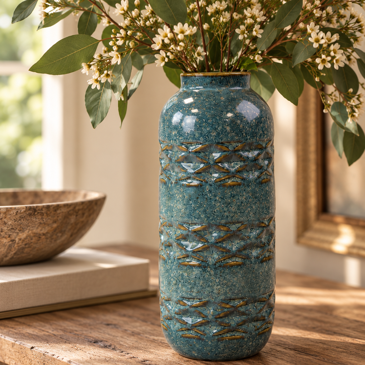 Close-up of a teal speckled ceramic vase with raised blue-grey and earthy brown detailing, filled with eucalyptus and delicate white flowers in a warm natural interior.