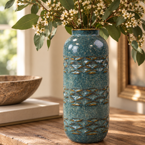 Close-up of a teal speckled ceramic vase with raised blue-grey and earthy brown detailing, filled with eucalyptus and delicate white flowers in a warm natural interior.