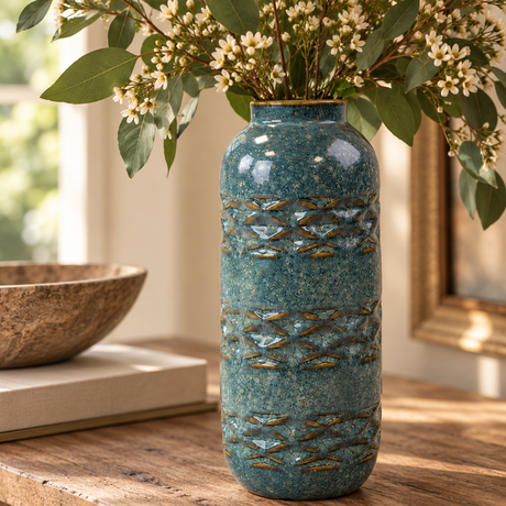 Close-up of a teal speckled ceramic vase with raised blue-grey and earthy brown detailing, filled with eucalyptus and delicate white flowers in a warm natural interior.