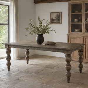 A rustic farmhouse dining table with distressed grey-brown wood and turned legs, styled in a bright dining room with a vase of greenery and plates on top.