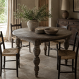 A round rustic wooden dining table with turned legs, surrounded by dark wood chairs with woven seats, in a softly lit dining room. A large vase with greenery and stacked plates sit on the tabletop.
