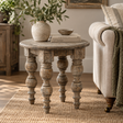 A distressed round wooden end table with turned legs, styled in a cosy living room next to a sofa with a throw blanket. The table is decorated with a textured ceramic vase containing green leaves and a stack of books.