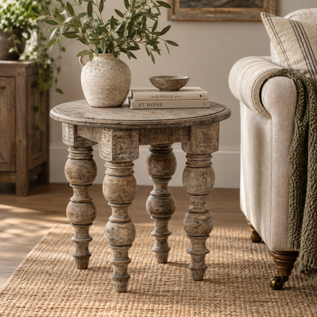 A distressed round wooden end table with turned legs, styled in a cosy living room next to a sofa with a throw blanket. The table is decorated with a textured ceramic vase containing green leaves and a stack of books.