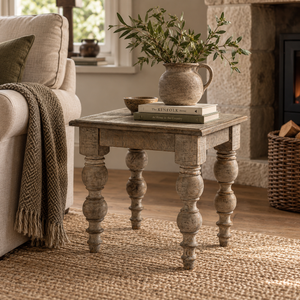 A rustic distressed wood end table styled in a cosy living room next to a beige armchair, holding a ceramic pitcher of greenery and books.