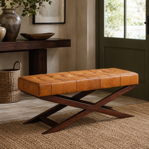 A tan leather tufted bench with a dark wood X-shaped base, styled in a warm, rustic room setting with a woven rug, wooden console table, and green door.