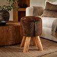 A round stool with a brown animal-hide textured top and four natural wood legs, styled in a cosy living room next to a white sofa and wooden coffee table.