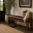 A dark brown tufted leather bench with a wooden frame in a styled entryway, featuring a woven straw tote bag, a cream throw blanket, and a pair of brown shoes.