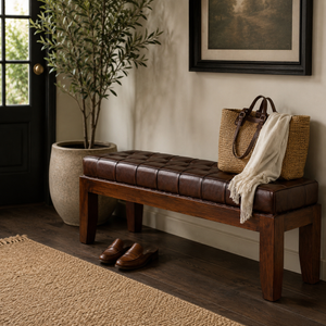 A dark brown tufted leather bench with a wooden frame in a styled entryway, featuring a woven straw tote bag, a cream throw blanket, and a pair of brown shoes.