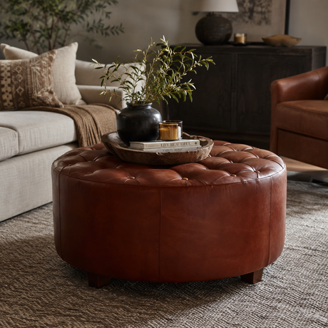 A round tufted leather ottoman in a warm reddish-brown colour, styled in a cosy living room setting with a tray, books, and a vase of greenery on top.