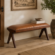 A brown leather bench with dark wood legs sits in a styled room corner with a framed print, books, a bowl, and a potted plant.