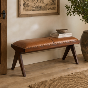 A brown leather bench with dark wood legs sits in a styled room corner with a framed print, books, a bowl, and a potted plant.