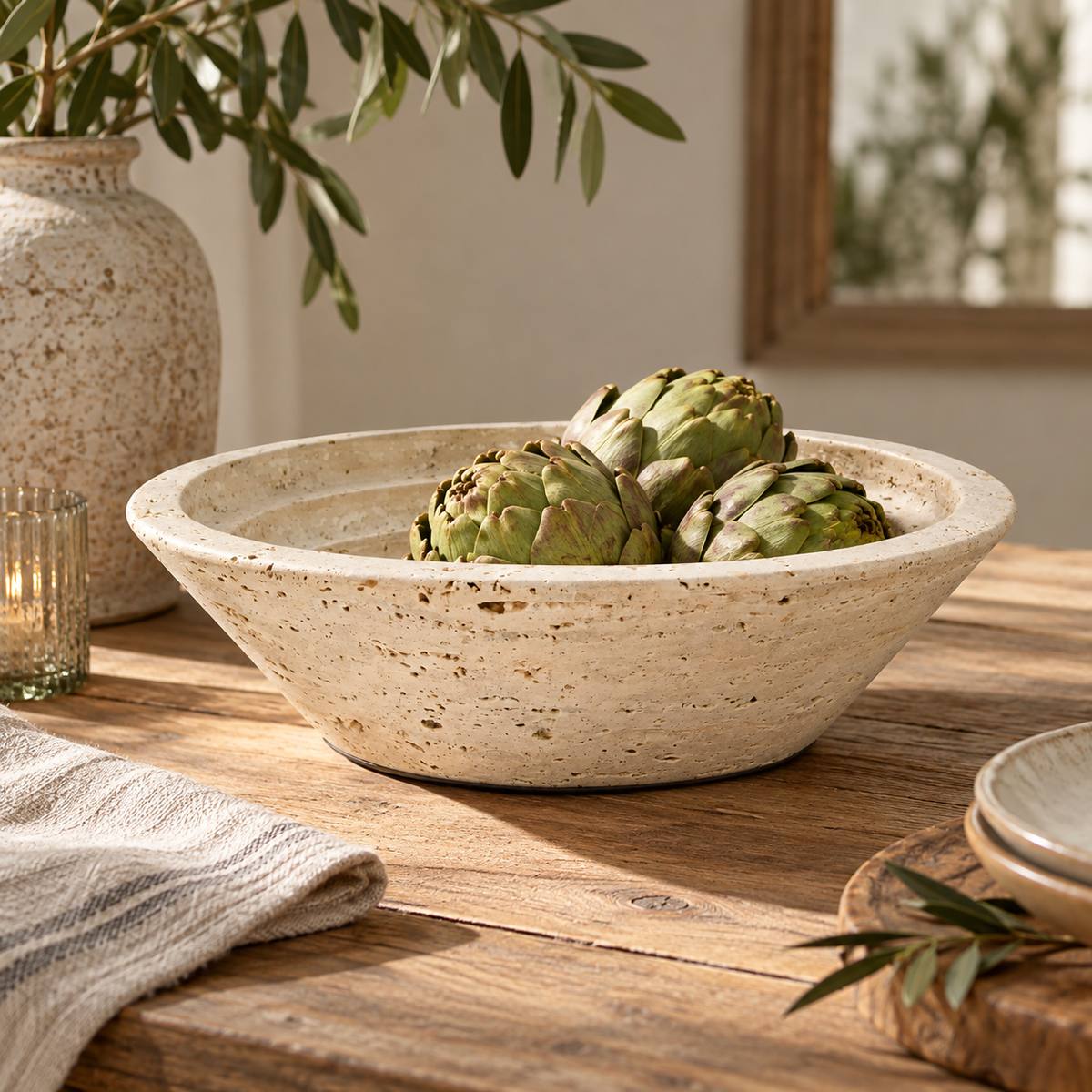 Tivoli Travertine Round Tapered Bowl, filled with green artichokes, set on a rustic wooden table with a striped cloth, a glass candle holder, and a large vase in the background.