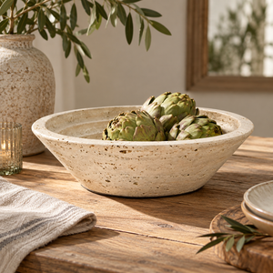 Tivoli Travertine Round Tapered Bowl, filled with green artichokes, set on a rustic wooden table with a striped cloth, a glass candle holder, and a large vase in the background.