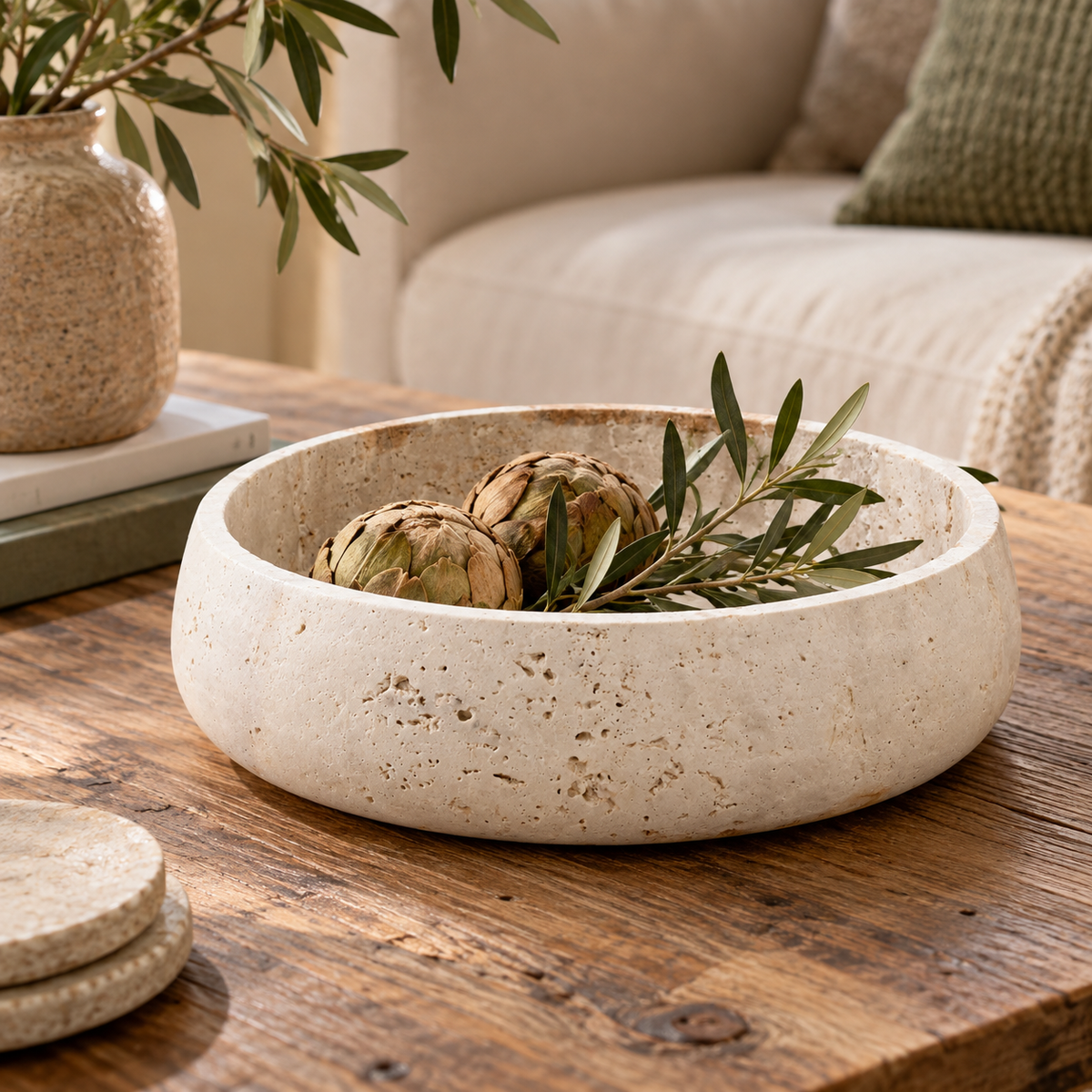 Tivoli Travertine Round Wide Rim Bowl in warm cream, styled on a rustic wooden table with artichokes and olive branches inside, with a matching vase and coasters in the background.