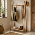 A tall wooden A-frame entryway coat rack with a slatted bottom shelf, shown in a styled home interior. The rack holds an olive green jacket, a beige tote bag, a beige scarf, a woven hat, and a pair of brown boots on the shelf.