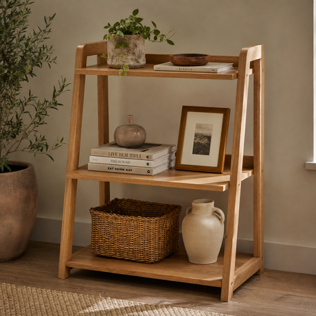 A three-tier wooden ladder shelf with open sides, styled with books, a potted plant, a vase, a framed picture, a wicker basket, and a ceramic jug in a warm, neutral room setting.