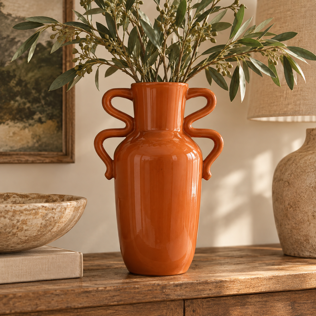 Tova Burnt Orange Tall Vase with Abstract Handles, filled with green olive branches, displayed on a wooden console table in a warm, styled room setting.