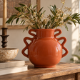 Tova Burnt Orange Wide Vase with Abstract Handles, styled with leafy branches and delicate white flowers on a rustic wooden table in a warm, neutral interior.
