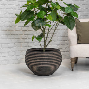 A potted houseplant with large green leaves in a dark ribbed round planter, placed indoors against a white brick wall with part of a light upholstered chair visible on the right.