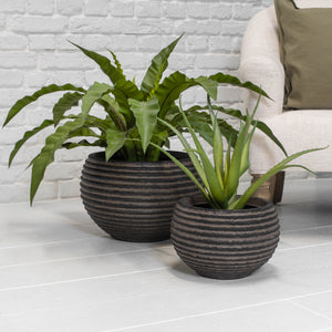 A pair of dark brown ribbed planters with green plants in next to a cream chair and with a background of a white brick wall