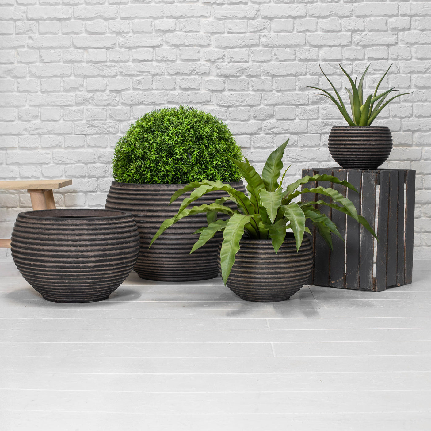 Four dark brown ribbed ceramic planters in various sizes, styled with green plants against a white brick wall and light grey floor.