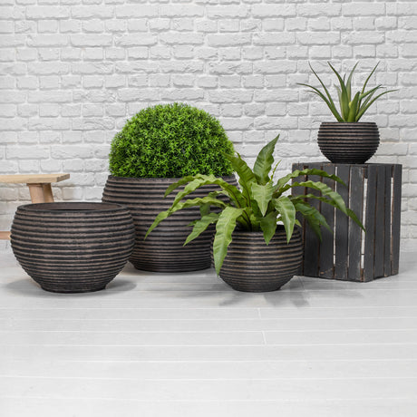 Four dark brown ribbed ceramic planters in various sizes, styled with green plants against a white brick wall and light grey floor.