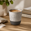 Speckled ceramic cup with a dark base on a wooden surface, with a plant and books in the background.