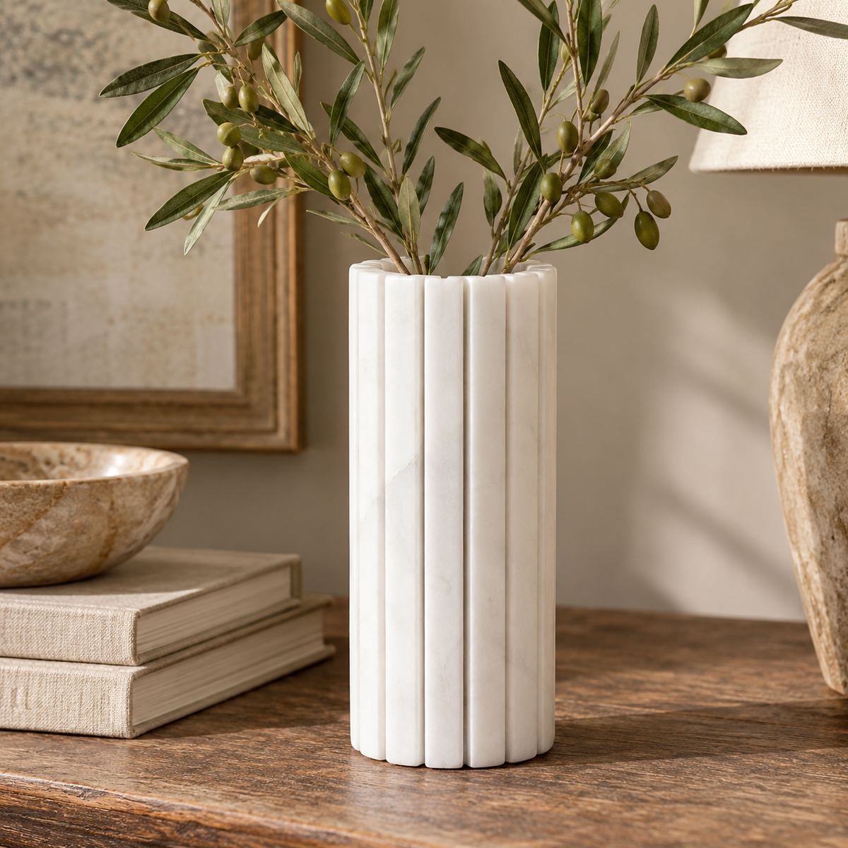 Vela White Marble Fluted Bud Vase, filled with olive branches, styled on a rustic wooden table in a warm, neutral interior with books and natural stone accents.