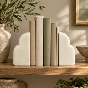 Vela White Marble Set of Two Bookends holding a row of linen and sage books on a rustic wooden shelf, with a stone vase of olive branches.