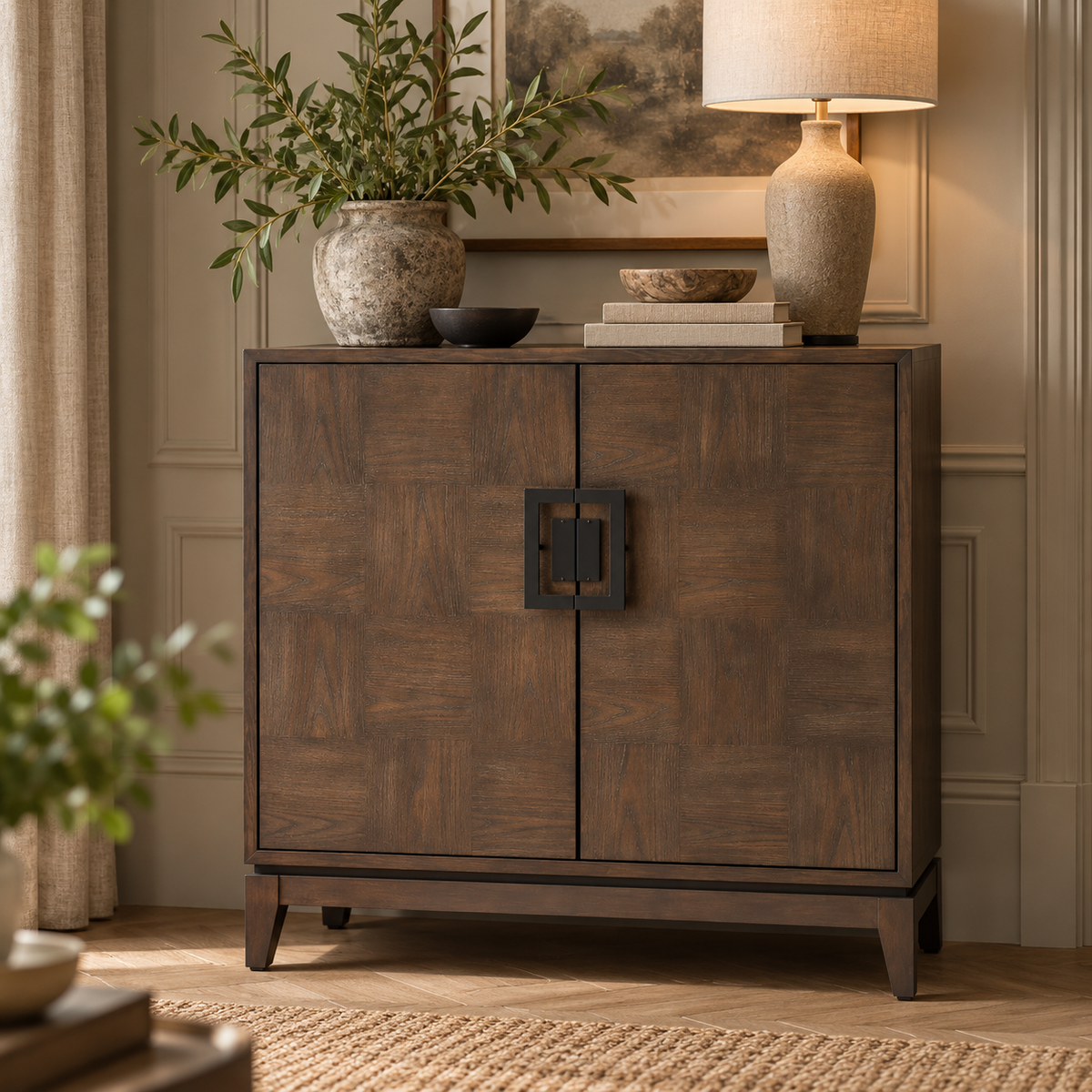 An antique grey wooden sideboard cabinet with a geometric black metal handle, styled in a warm, elegant room with a lamp, potted plant, and books on top.