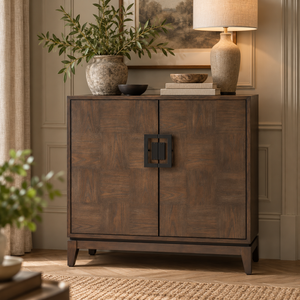 An antique grey wooden sideboard cabinet with a geometric black metal handle, styled in a warm, elegant room with a lamp, potted plant, and books on top.