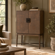 An antique grey wooden cabinet with two doors and a black metal handle, styled in a warm, neutral-toned living room next to a window and potted plants.