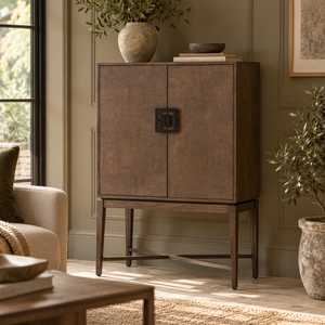 An antique grey wooden cabinet with two doors and a black metal handle, styled in a warm, neutral-toned living room next to a window and potted plants.