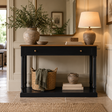 A black console table with a light wood top, styled in a warm, elegant room. On top are a large textured ceramic vase with greenery and a matching ceramic table lamp.