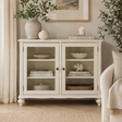 A white wooden sideboard cabinet with two glass-front doors and open shelves, styled in a bright living room with books, vases, and plants.