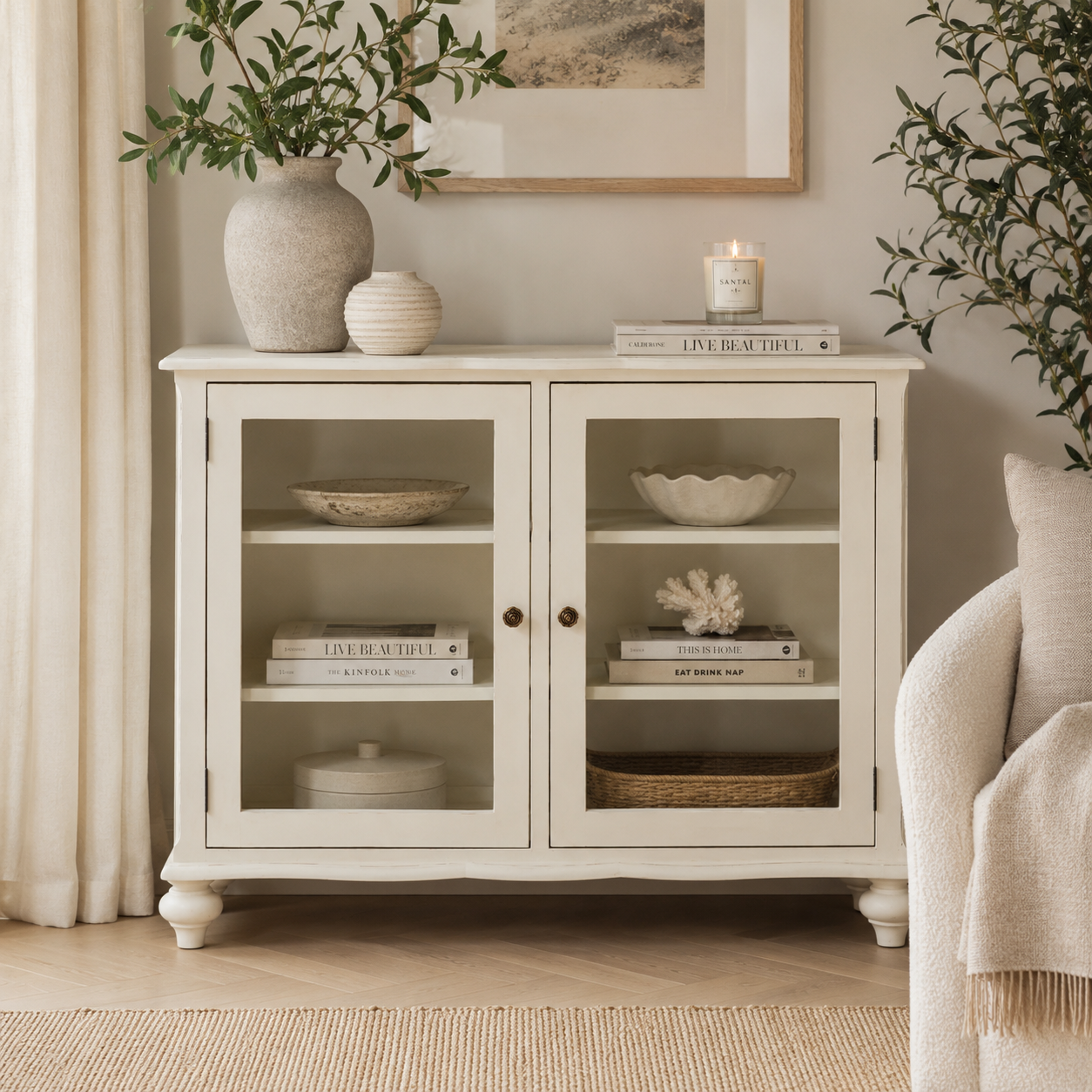 A white wooden sideboard cabinet with two glass-front doors and open shelves, styled in a bright living room with books, vases, and plants.